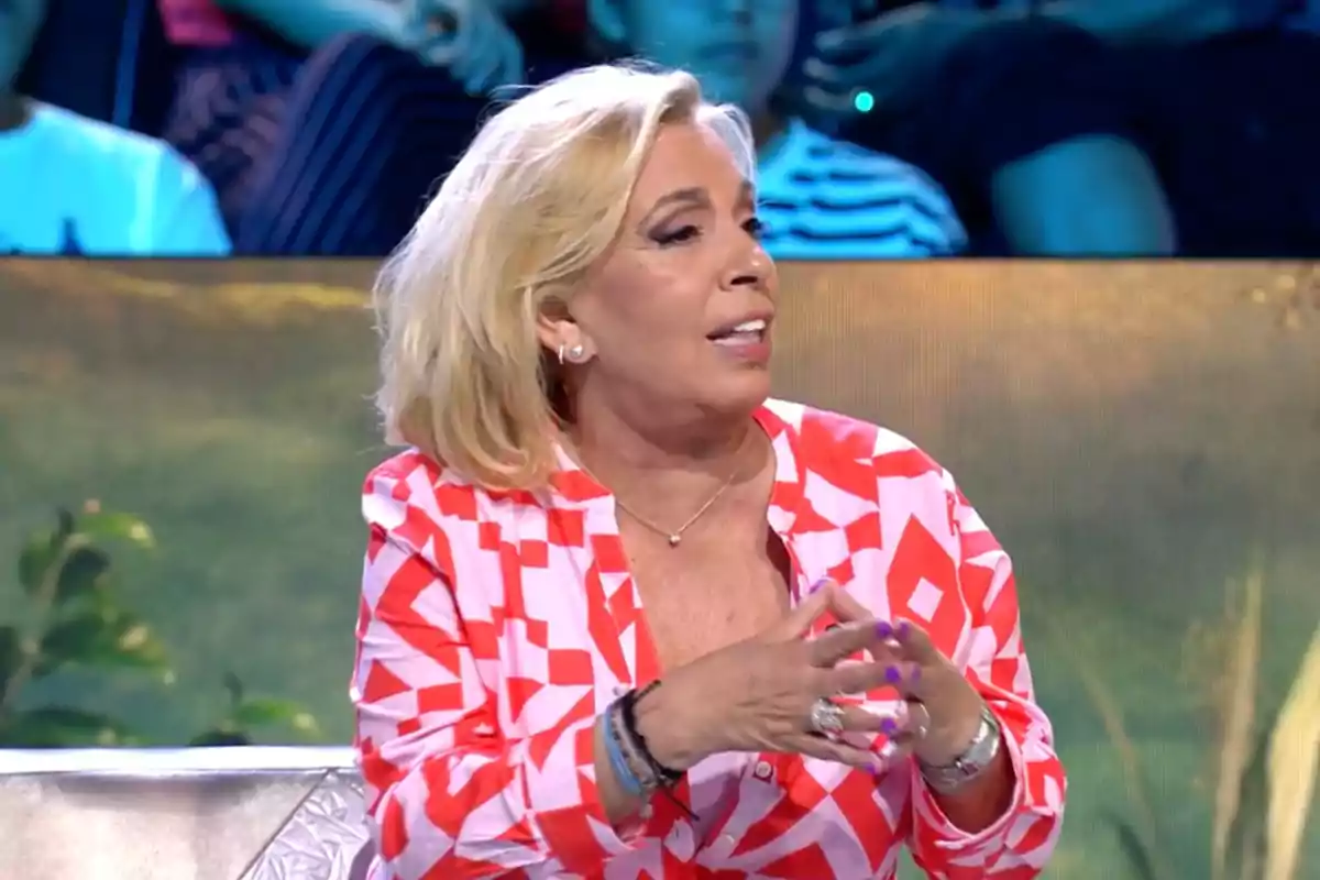 Carmen Borrego with blonde hair and a red and white geometric patterned jersey is speaking on 'Supervivientes' with a blurred audience background. Carmen Borrego with blonde hair and a red and white geometric patterned jersey is speaking on 'Supervivientes' with a blurred audience background.