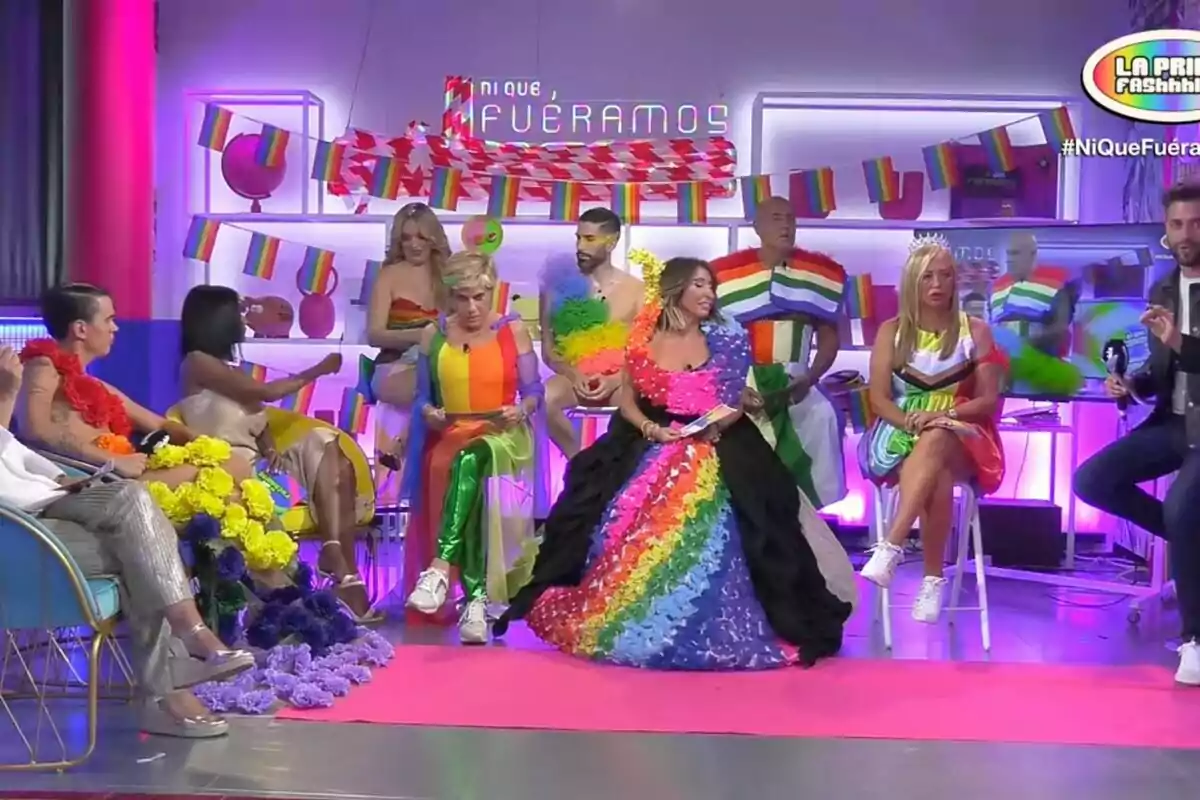 Un grupo de personas vestidas con ropa colorida y temática del orgullo LGBT+ están sentadas en un set de televisión decorado con banderas de arcoíris y luces de colores.