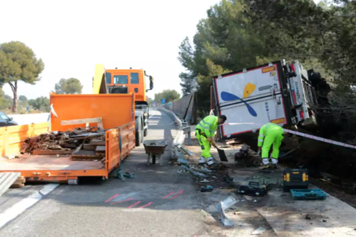 Imatge del punt de l'AP-7, al Vendrell (Tarragona), on apareix un camió bolcat fora de la via i dues persones fent la seva feina el dia posterior a l'accident
