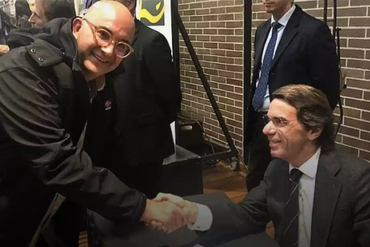Al costat esquerre, Xavier Rius una mica ajupit, somrient i donant-li la mà a José María Aznar assegut i somrient