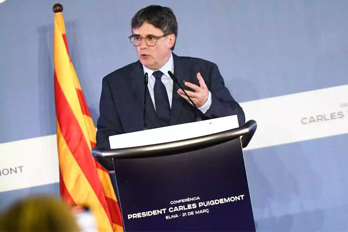 Carles Puigdemont parlant des d'un faristol de color blau amb una bandera catalana de fons