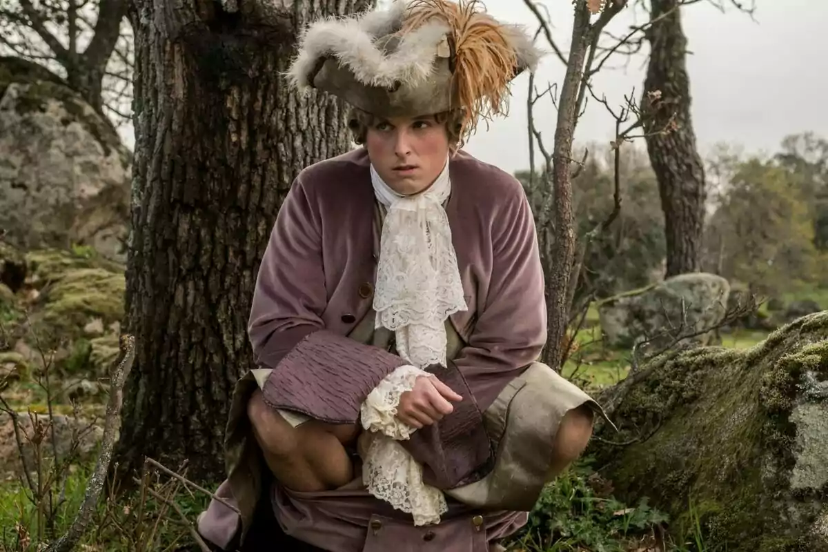 Carlos Scholz vestido con ropa de época, incluyendo un sombrero con plumas, está agachado junto a un árbol en un entorno natural en La Vida Breve.