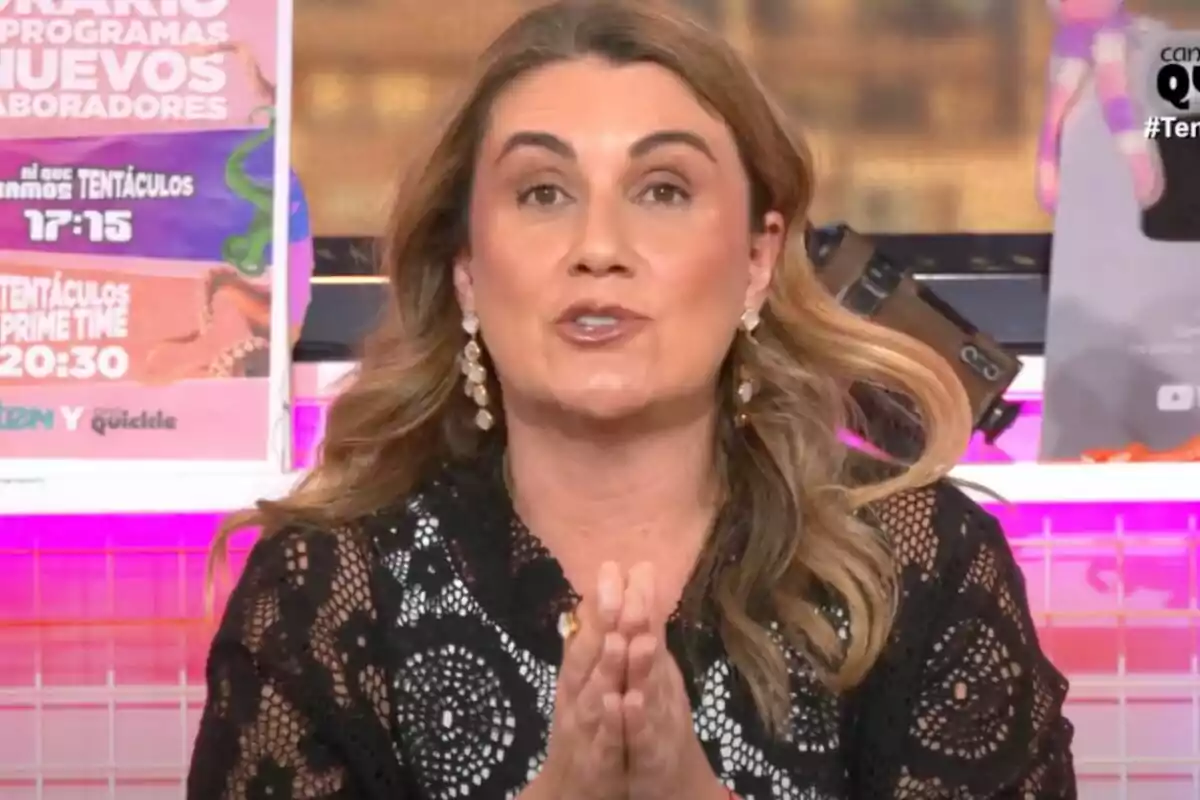 Carlota Corredera amb cabell llarg i solt, vestida amb una brusa negra de puntes, està parlant davant de la càmera a 'Tentáculos', amb cartells acolorits al fons.