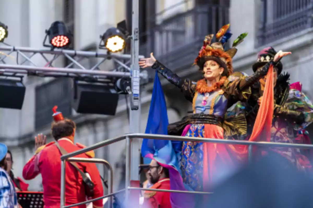 Una persona vestida con un colorido disfraz y sombrero decorado está de pie en una plataforma elevada, rodeada de luces y otras personas también disfrazadas, en lo que parece ser un evento festivo o desfile.