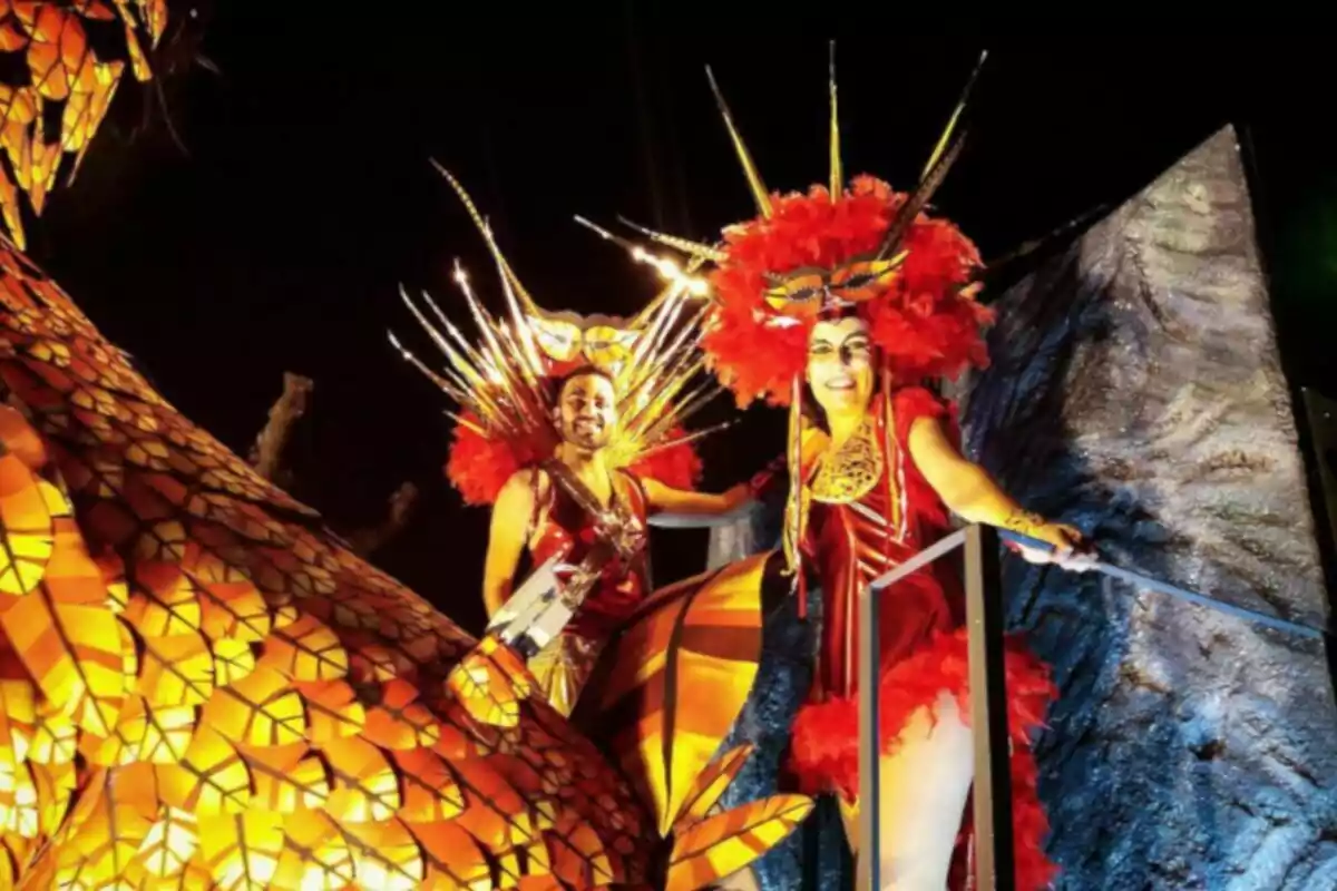 Personas con trajes coloridos y tocados elaborados posan en un carro alegórico iluminado durante un desfile nocturno.