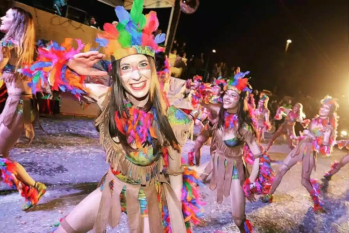 Grupo de personas disfrazadas con plumas coloridas bailando en un desfile nocturno.