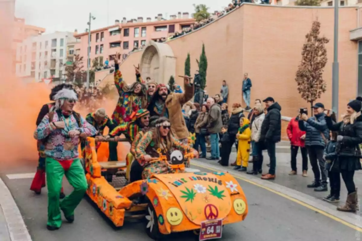 Un grup de persones disfressades amb roba acolorida i temàtica hippie participa en una desfilada de carrer amb un vehicle decorat, mentre el públic observa i fa fotos.