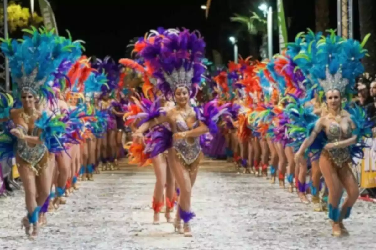 A group of dancers in colorful costumes and feathers parade at a nighttime event.