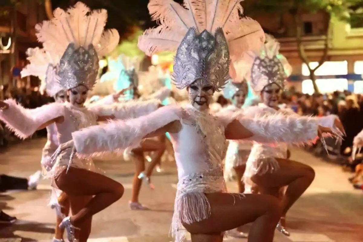 Grupo de bailarinas con trajes brillantes y plumas realizando una coreografía en un desfile nocturno.