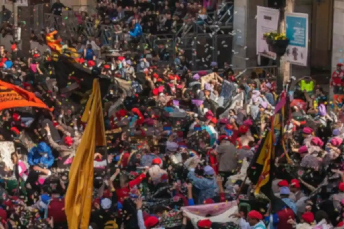 A crowd of people with red hats and scarves participates in an outdoor festive event, surrounded by confetti and colorful flags.