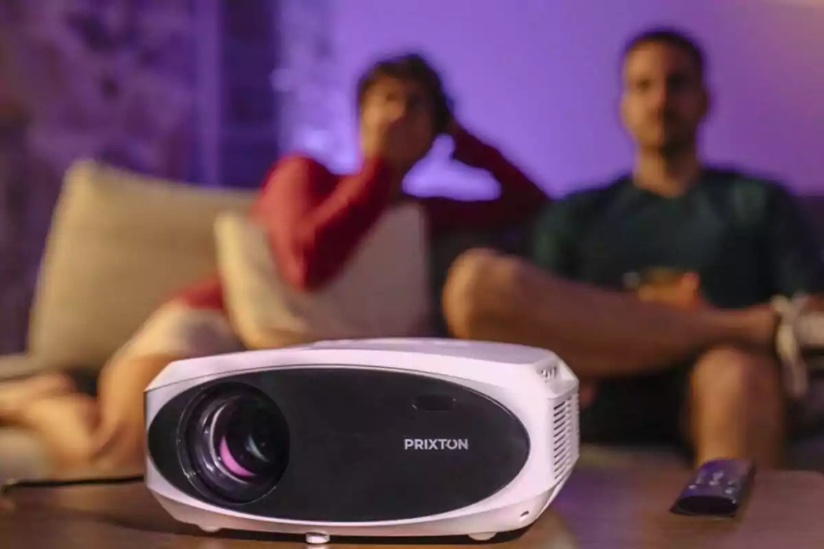 A Prixton projector in the foreground with two blurred people in the background sitting on a sofa. A Prixton projector in the foreground with two blurred people in the background sitting on a sofa.