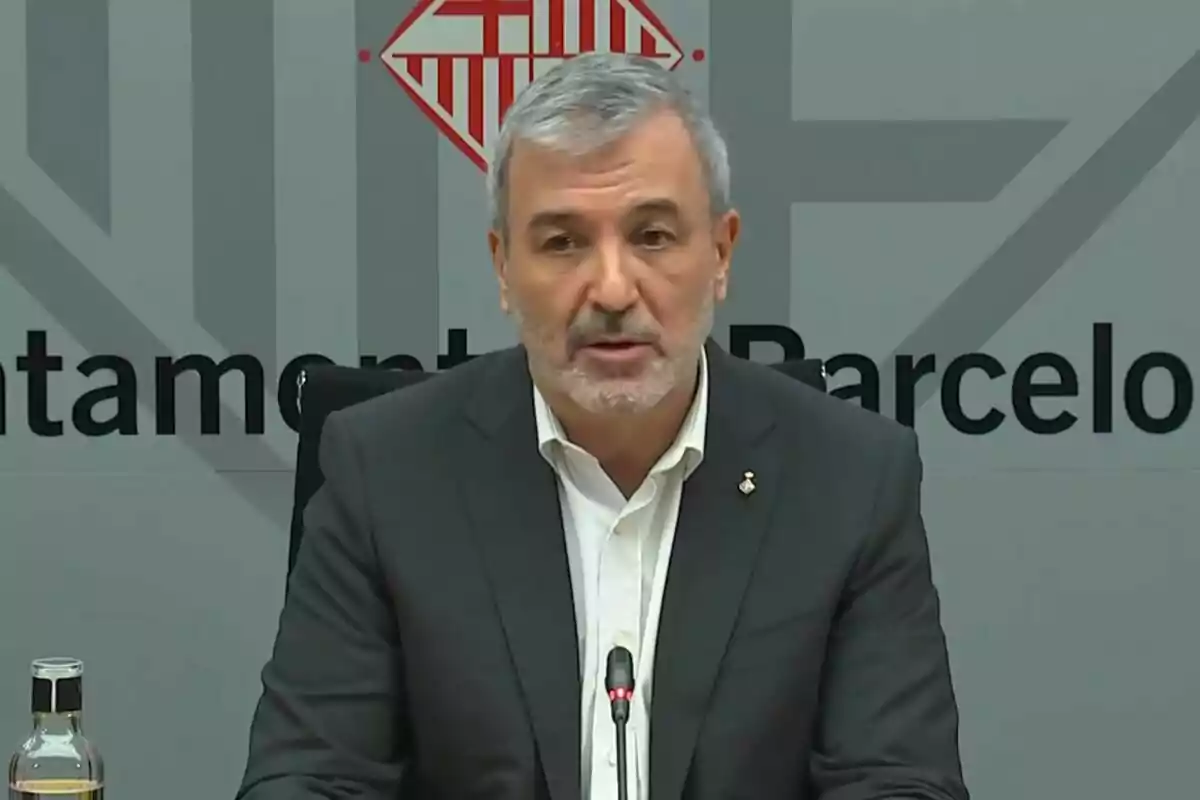 Gray-haired man with a beard speaking at a press conference with a background featuring the Barcelona City Hall logo.