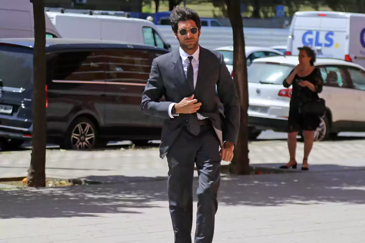 Cayetano Rivera in a suit and sunglasses walks down the sidewalk in an urban setting with parked vehicles in the background. Cayetano Rivera in a suit and sunglasses walks down the sidewalk in an urban setting with parked vehicles in the background.