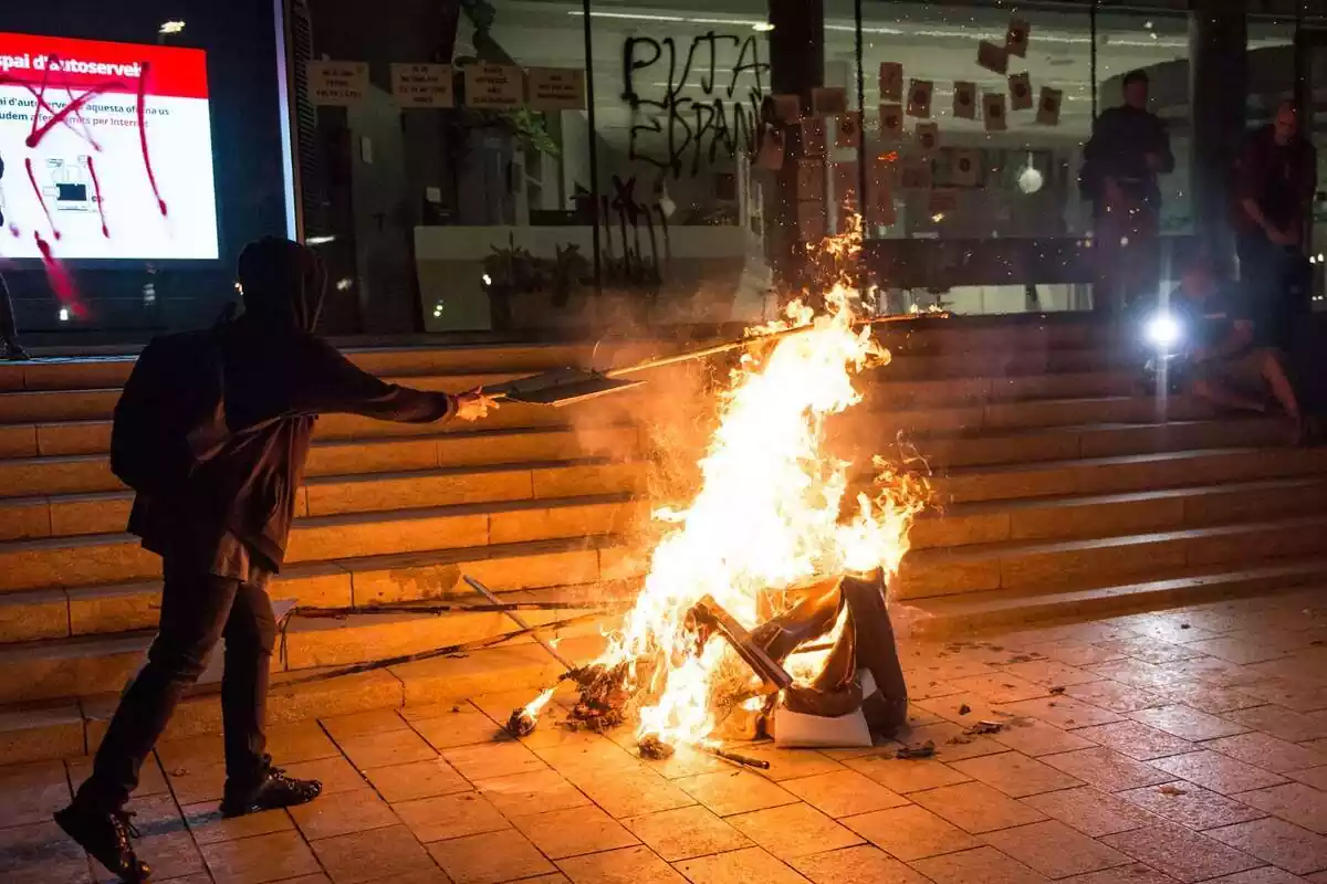 Pla general d'un manifestant independentista vestit de negre i encaputxat llançant un objecte a una foguera situada davant d'un edifici que té un grafiti que posa 'Puta Espanya' i una estelada