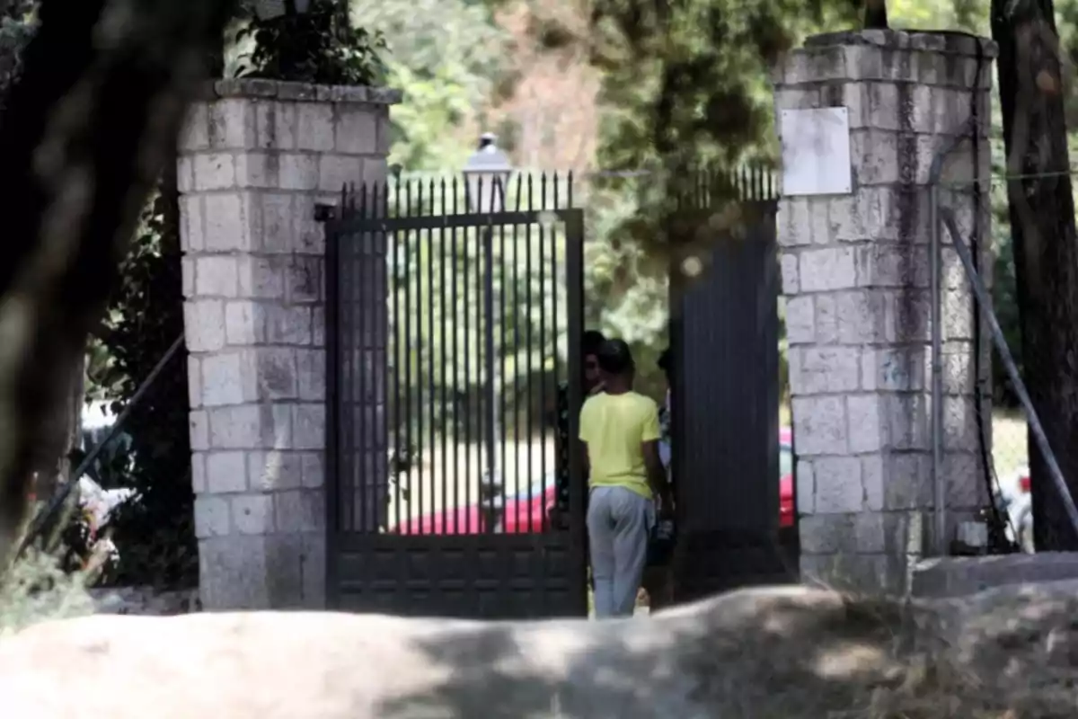 Una persona amb samarreta groga i pantalons grisos està dreta davant d'una porta de ferro entre dos pilars de pedra en un entorn arbrat.