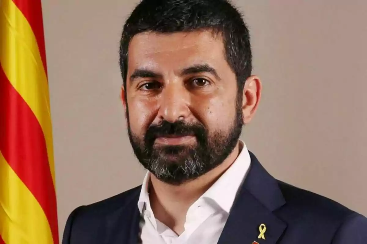 A man with a beard and short hair, dressed in a dark suit and white jersey, wears a yellow ribbon on his lapel, with a flag in the background. A man with a beard and short hair, dressed in a dark suit and white jersey, wears a yellow ribbon on his lapel, with a flag in the background.