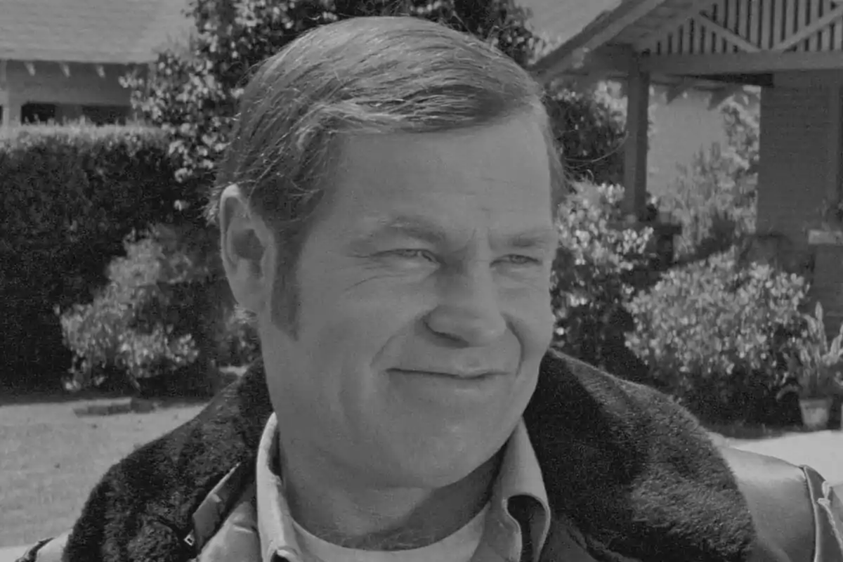 Fotografía en blanco y negro de Charles Cyphers sonriente con patillas y cabello corto, de pie al aire libre frente a una casa y vegetación en la película Halloween