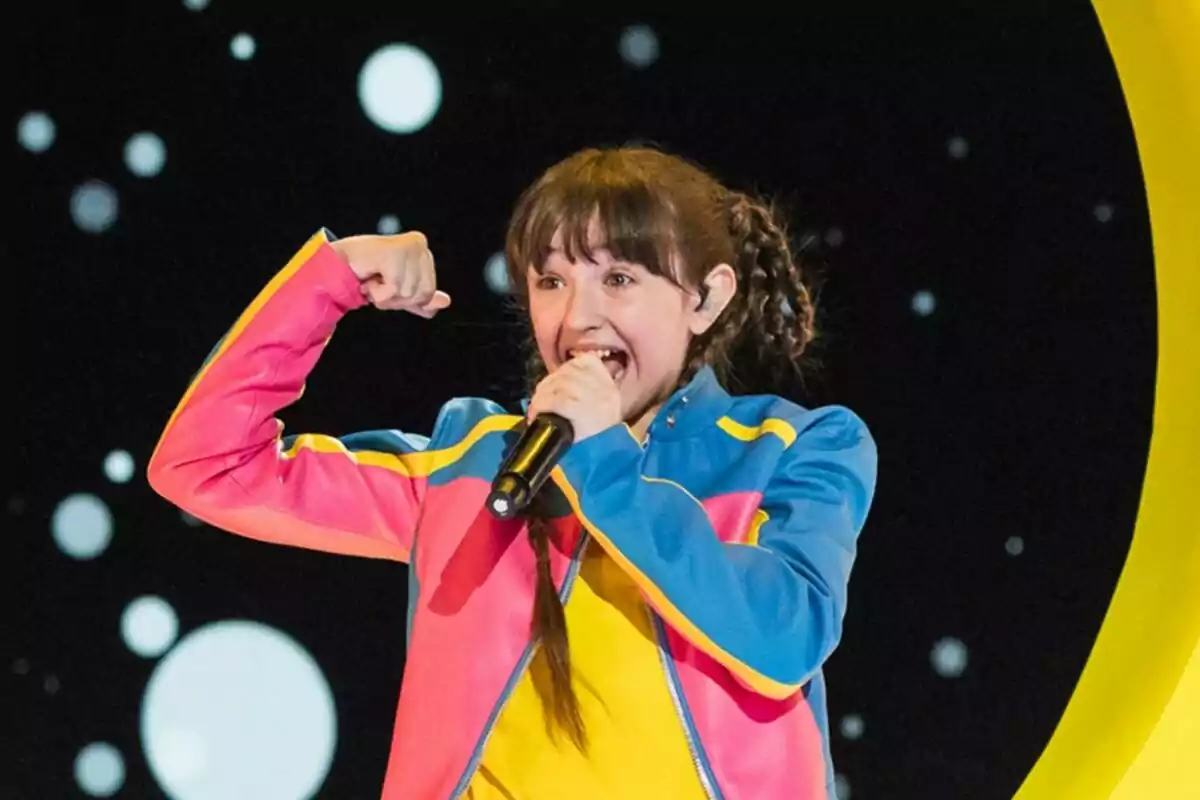 Chloe Delarosa amb jaqueta acolorida canta emocionada en un escenari amb fons de cercles blancs a Eurovisió Junior.