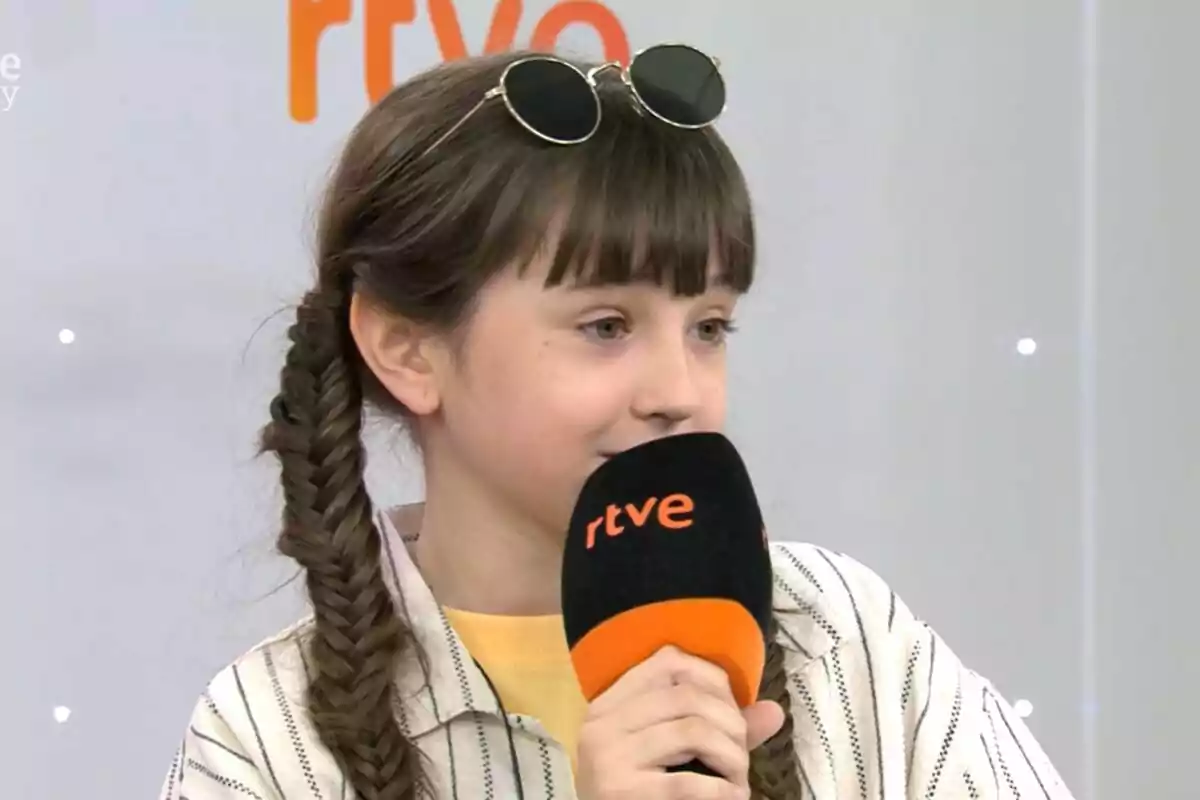 Fotografía de Chloe DelaRosa con trenzas y gafas de sol en la cabeza sostiene un micrófono de RTVE mientras habla en la presentación de Eurovisión Junior