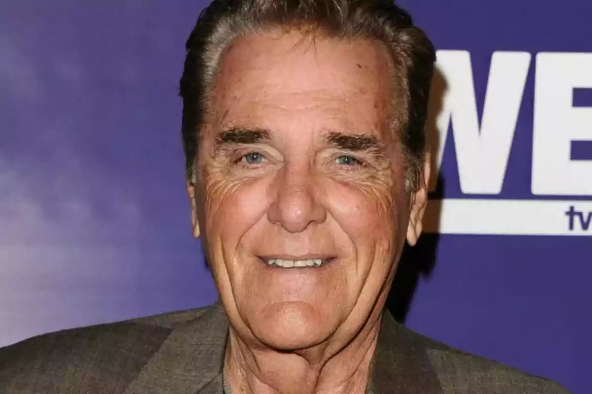 Chuck Woolery major amb cabell canós i ulls blaus somriu davant d'un fons morat.