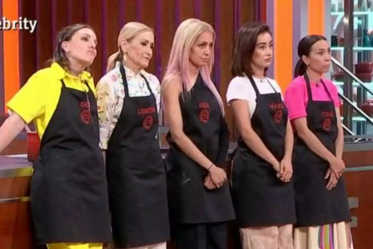 Inés Hernand, Cristina Cifuentes, Hiba Abouk, Marina Rivers i Itziar Miranda amb davantals negres de MasterChef Celebrity estan drets en una cuina de televisió.