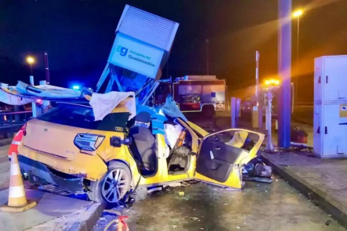 A yellow car crashed into a structure on a road at night with emergency lights and a fire truck in the background.