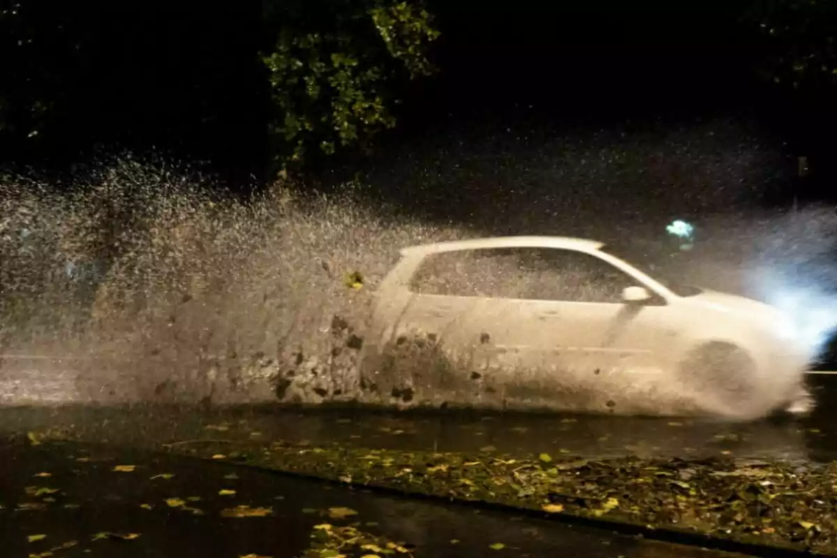 Un cotxe blanc esquitxant aigua mentre passa per un bassal en un carrer fosc. Un cotxe blanc esquitxant aigua mentre passa per un bassal en un carrer fosc.