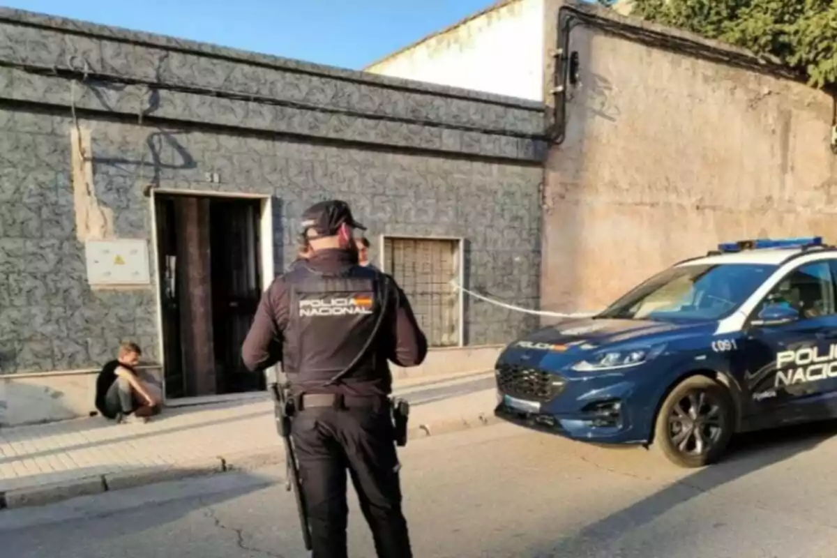 Un agent de la Policia Nacional està dret davant d'un edifici, mentre un cotxe patrulla està estacionat a prop i una persona està asseguda a la vorera.