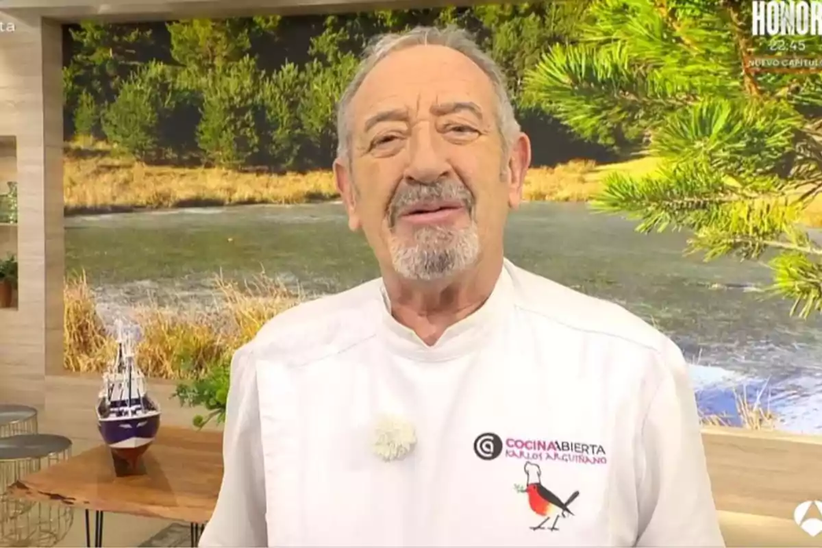 Karlos Arguiñano amb barba i bigoti està dret en un estudi de televisió amb un fons de paisatge natural i porta una jaqueta de xef blanca amb un logotip brodat. Captura de 'Cuina Oberta'.