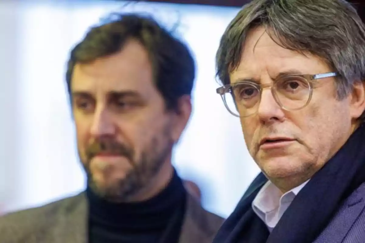 Two men with serious expressions, one in the foreground with glasses and the other blurred in the background.