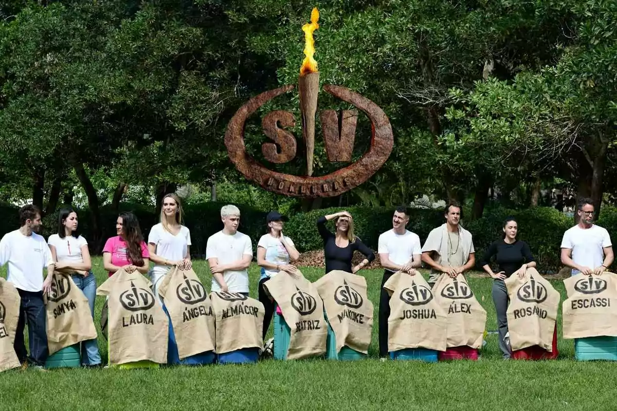 Un grupo de personas formado por Makoke, Pelayo Díaz, Álvaro Escassi, Almácor, Beatriz Rico, Laura Cuevas, Joshua Velázquez, Álex Adrover, Rosario Matew, Samya y Ángela Ponce de pie en un área verde sosteniendo sacos con nombres y un logotipo grande de SV (Supervivientes) en el fondo.