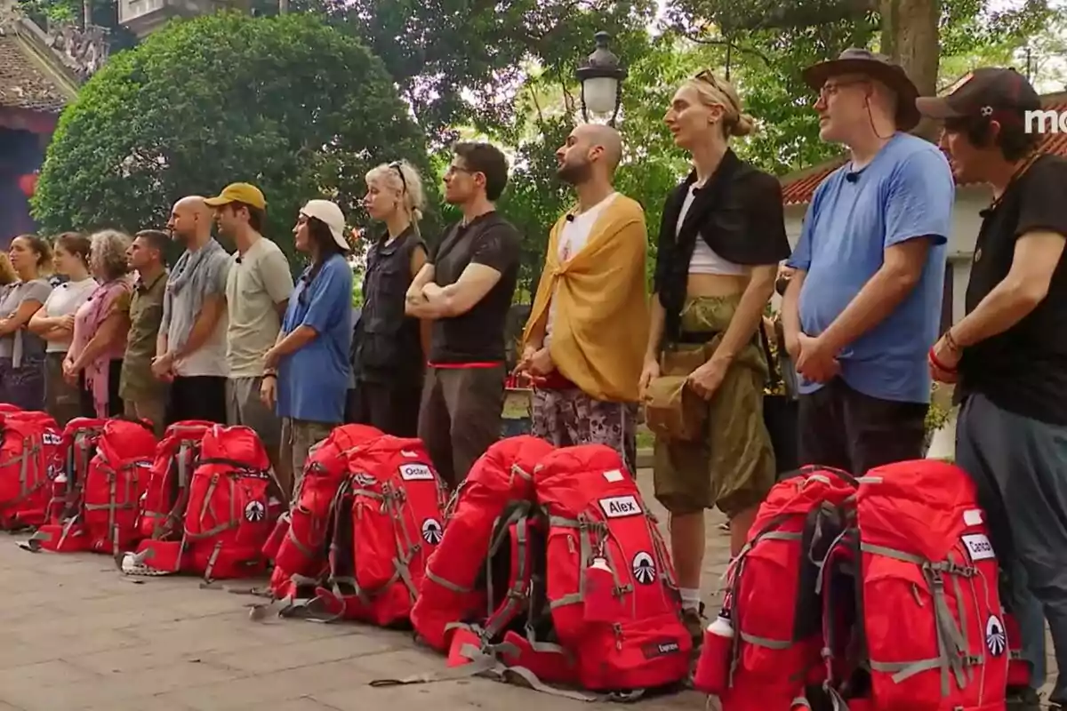 Un grup de concursants de peu en fila al costat de motxilles vermelles en un entorn a l'aire lliure amb arbres i un edifici al fons a la promo de Pekin Express.