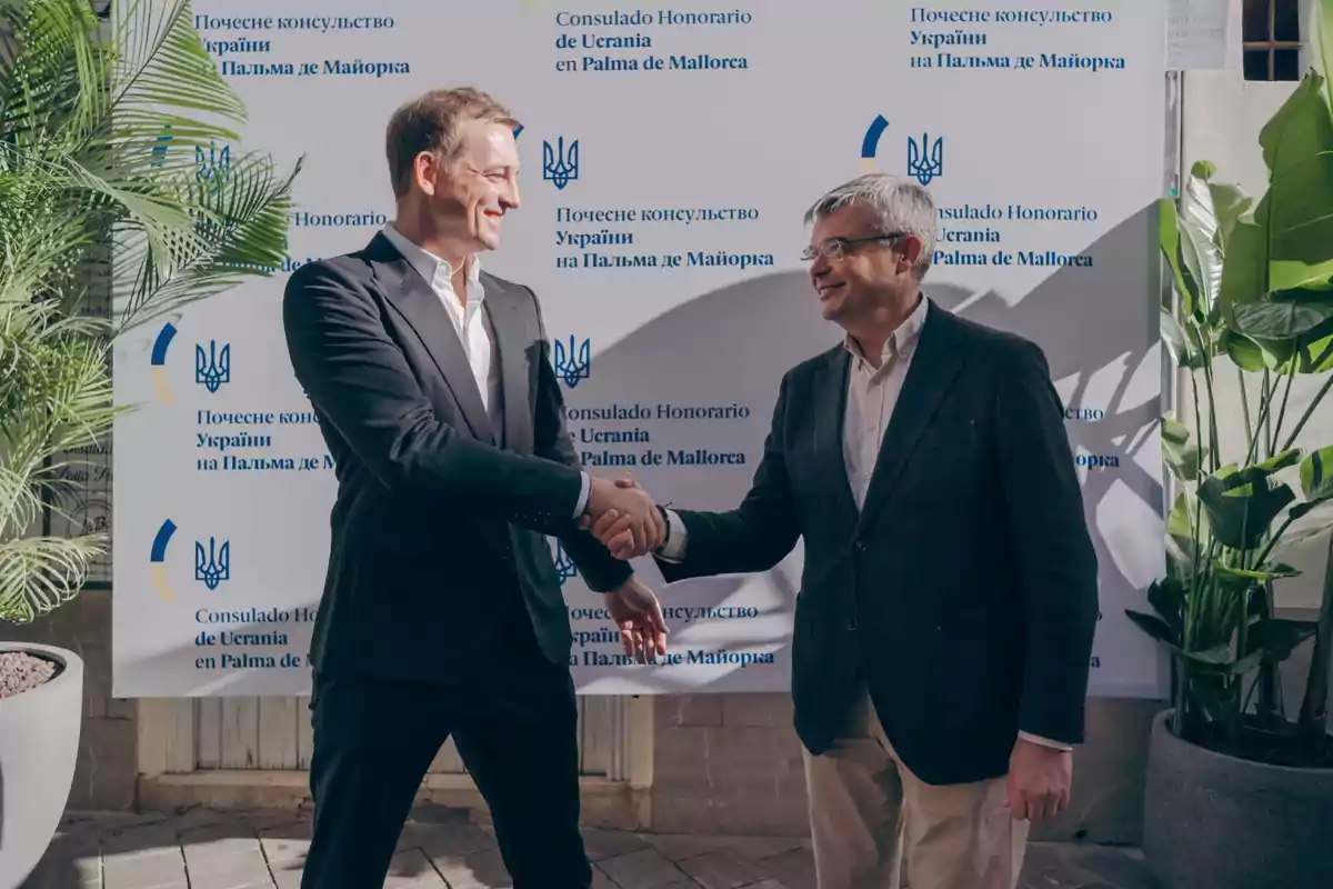 Dos hombres estrechándose la mano frente a un cartel del Consulado Honorario de Ucrania en Palma de Mallorca.