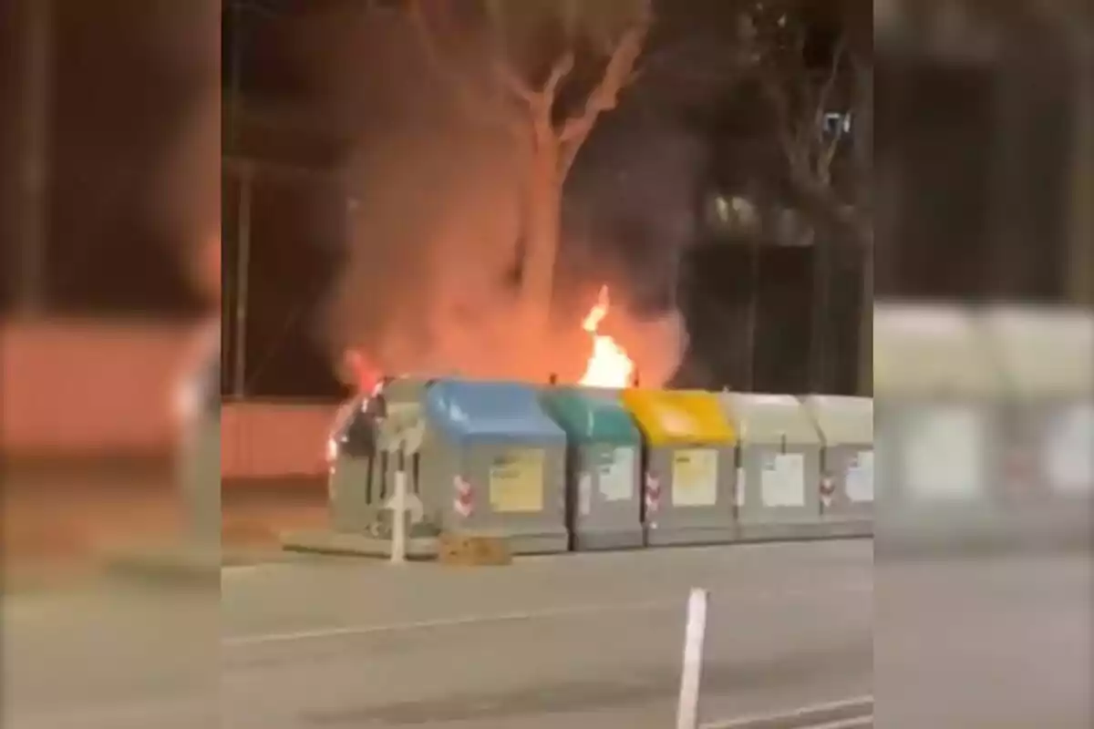 Contenedores de reciclaje en la calle con un incendio en el fondo.