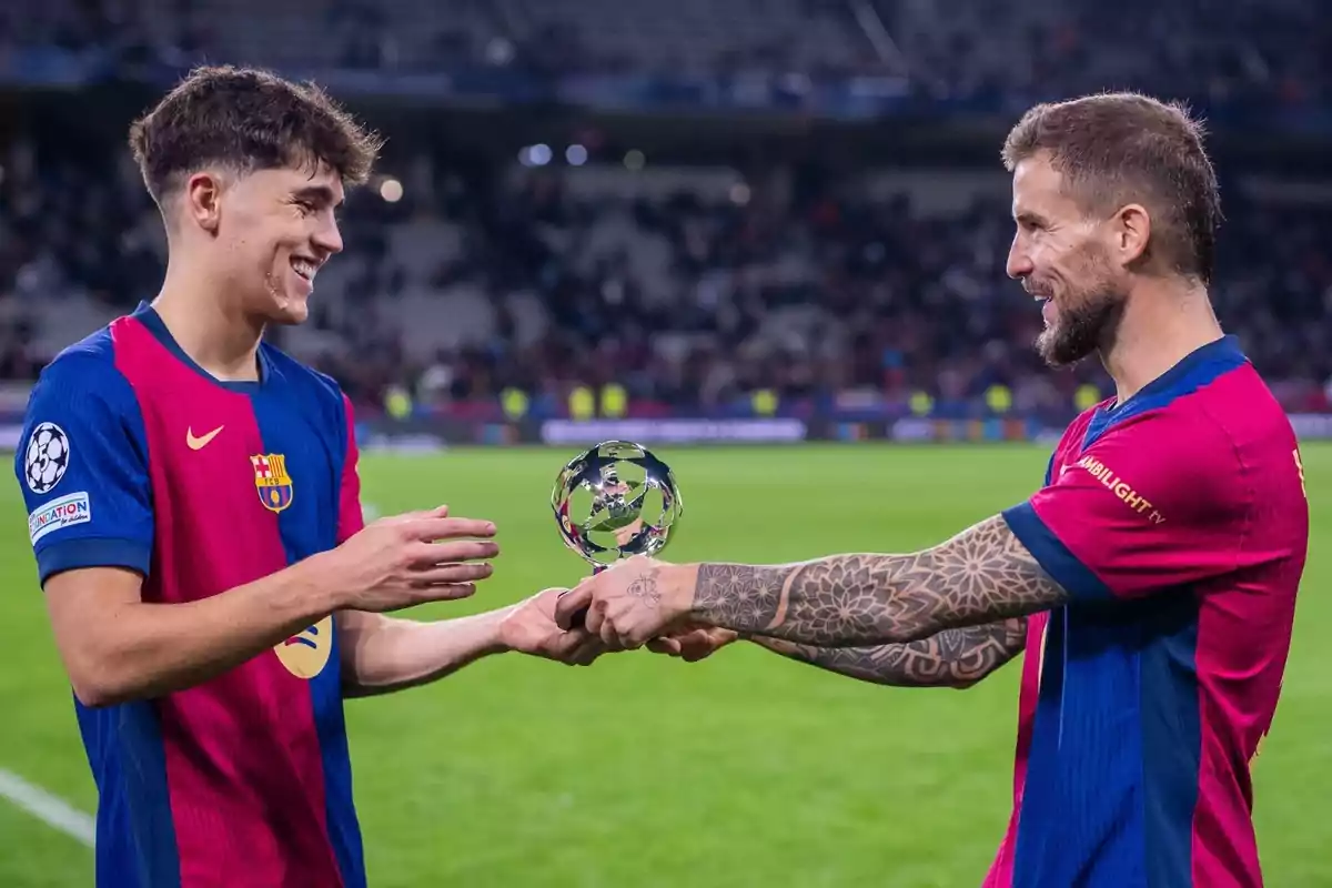 Two Barcelona players smile as one hands a trophy to the other on the field. Two Barcelona players smile as one hands a trophy to the other on the field.