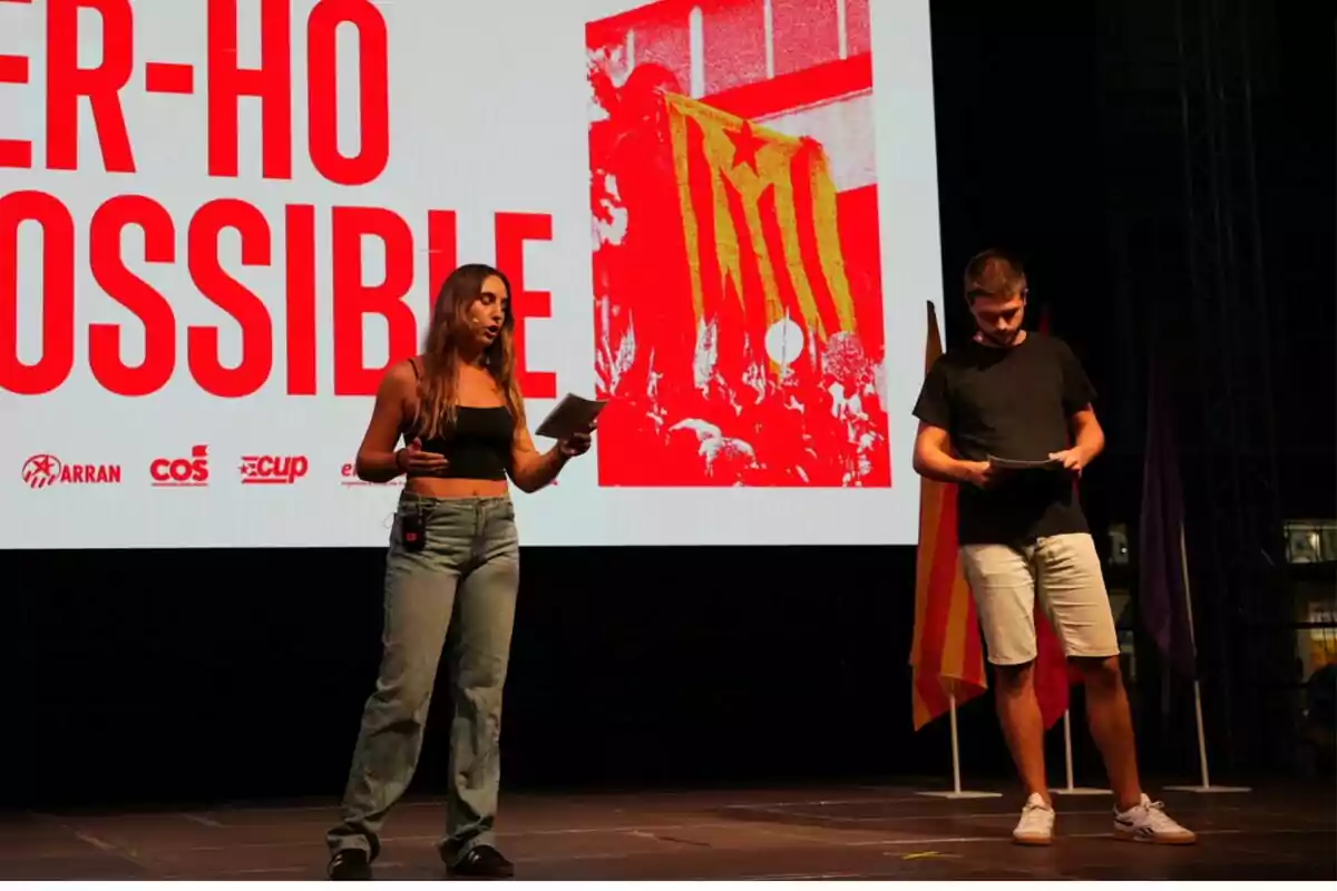 Dues persones en un escenari amb una pantalla de fons que mostra un missatge en català i diverses banderes.