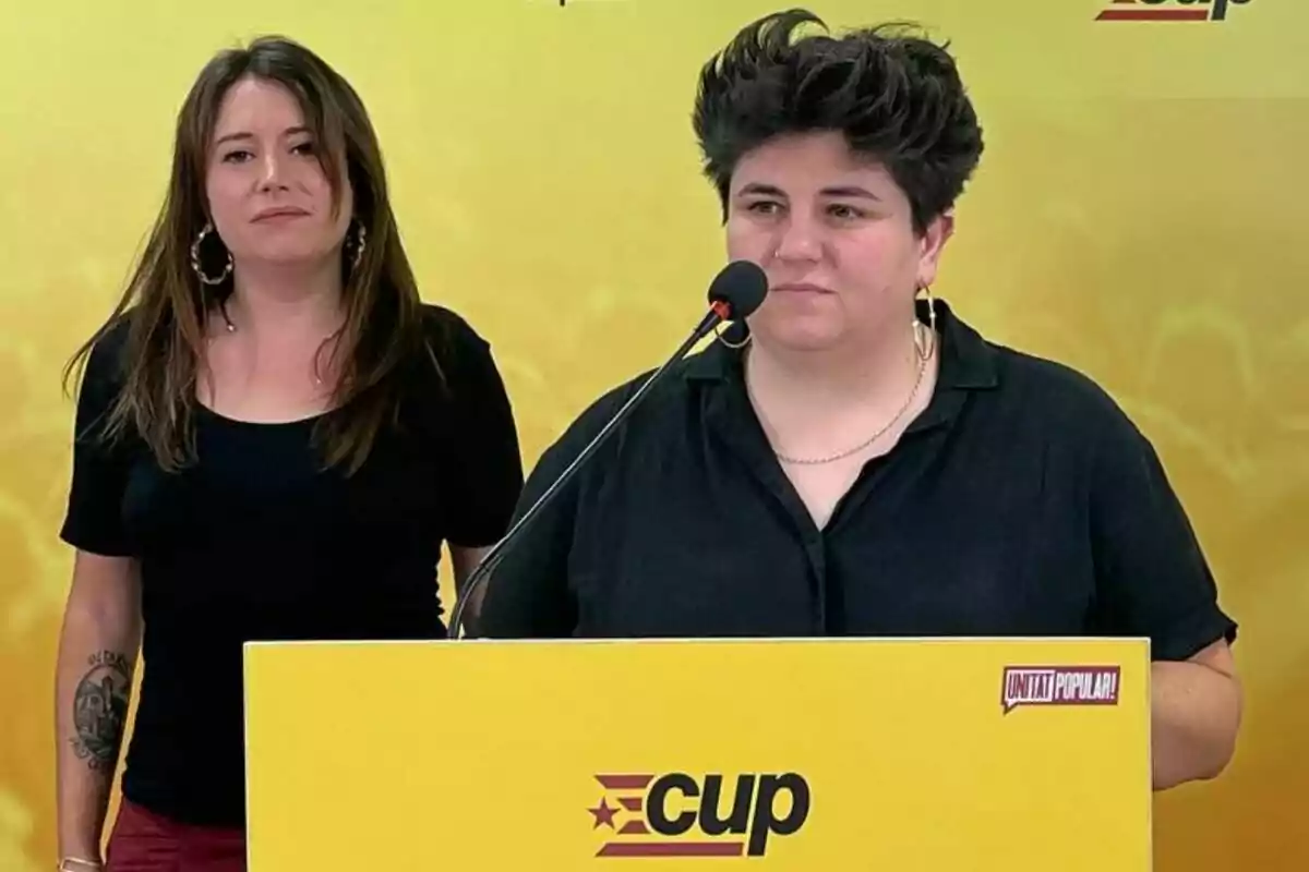 Adriana Llena y Laure Vega están de pie frente a un fondo amarillo con un logotipo de "CUP" en un podio, una de ellas habla por un micrófono.