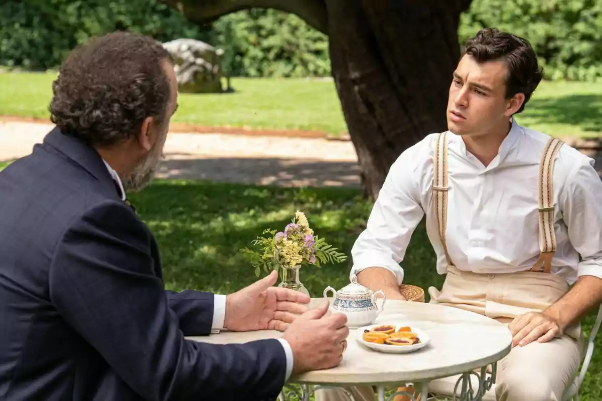 Curro i Alonso conversen en una taula a l'aire lliure en un jardí, un porta tirants i una camisa blanca mentre l'altre vesteix un vestit fosc, a la sèrie La Promesa.