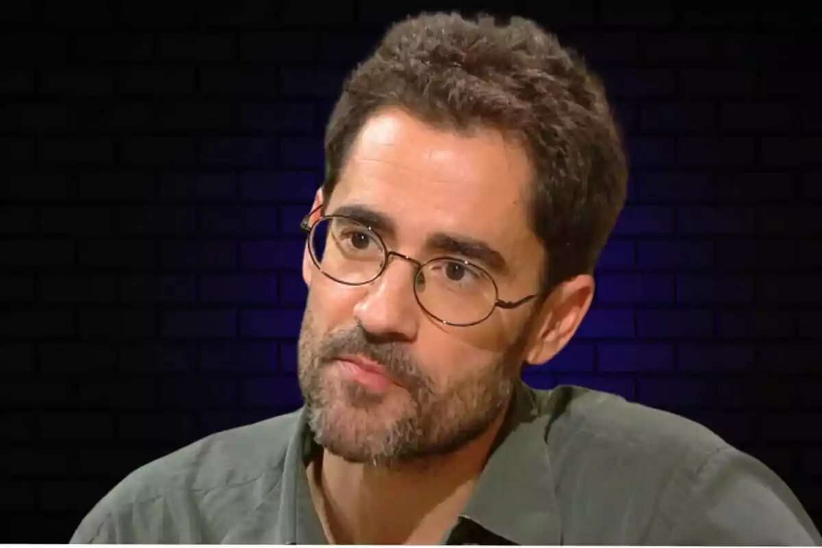 Un hombre con gafas y barba, vistiendo una camisa verde, con un fondo de ladrillos oscuros.