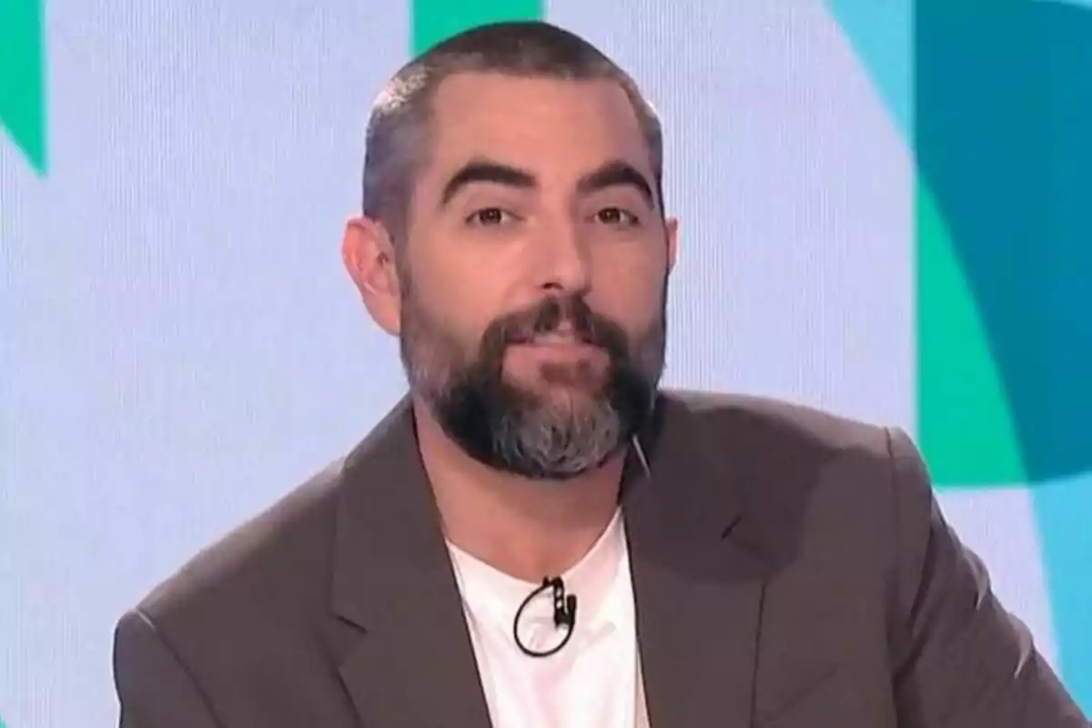 Dani Mateo amb barba i cabell curt porta un vestit marró i una samarreta blanca mentre és a Zapeando.