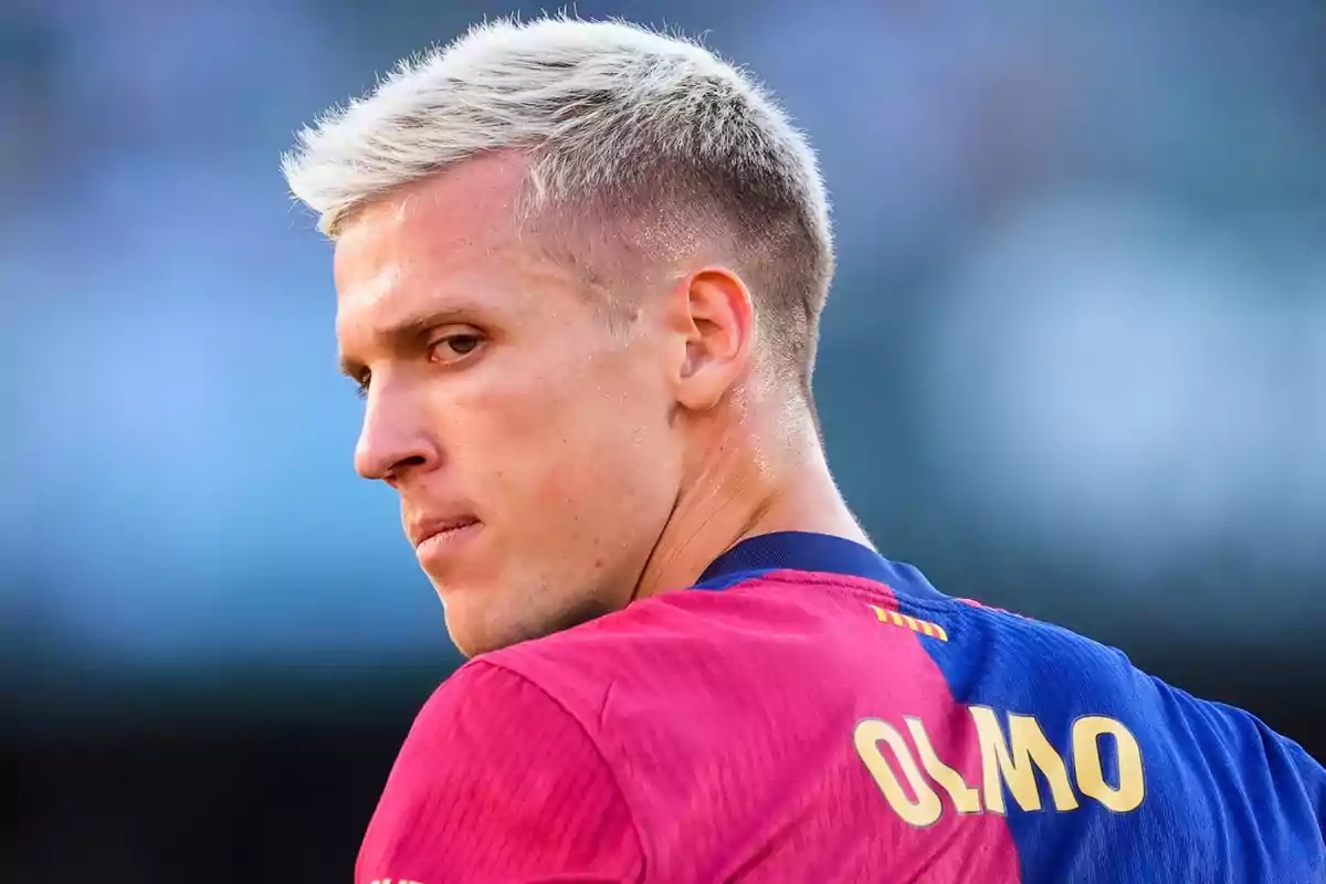 Un jugador de fútbol con cabello rubio y camiseta azul y roja mira hacia un lado.