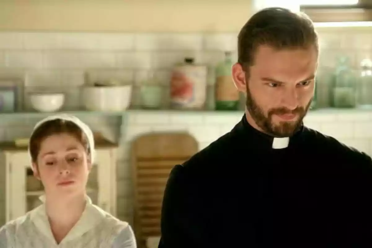 Imatge de l’actor Daniel Schröder vestit de capellà i una dona amb còfia blanca són en una cuina antiga amb prestatges i utensilis al fons. Imatge de l’actor Daniel Schröder vestit de capellà i una dona amb còfia blanca són en una cuina antiga amb prestatges i utensilis al fons.