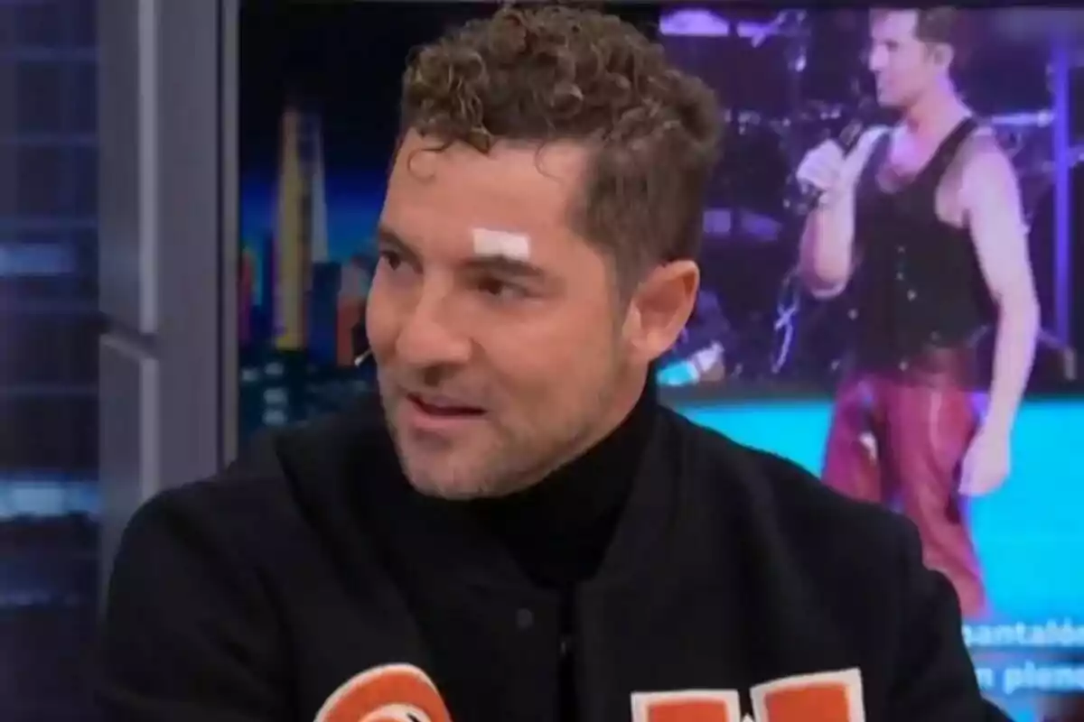 David Bisbal amb cabells arrissats i una cureta a la cella està parlant a El Hormiguero amb una imatge de fons d'una persona cantant.