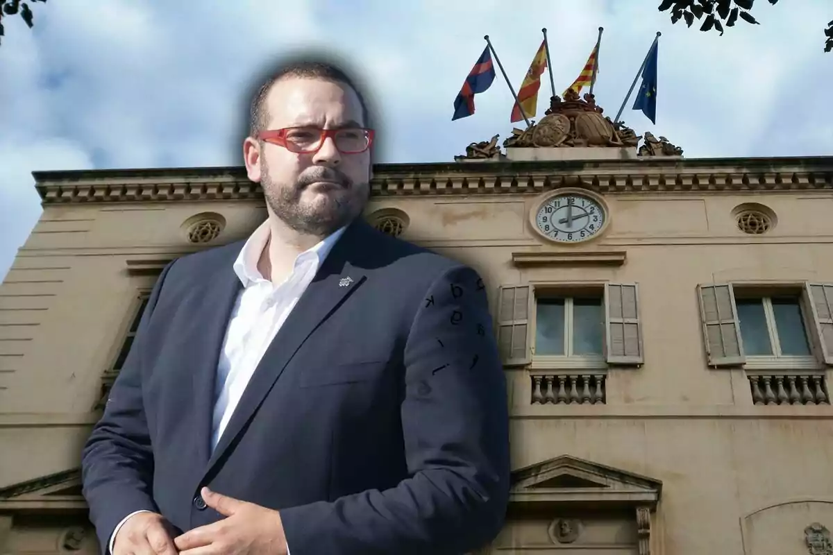 Home amb ulleres vermelles i barba davant d'un edifici amb un rellotge i diverses banderes onejant a la part superior.