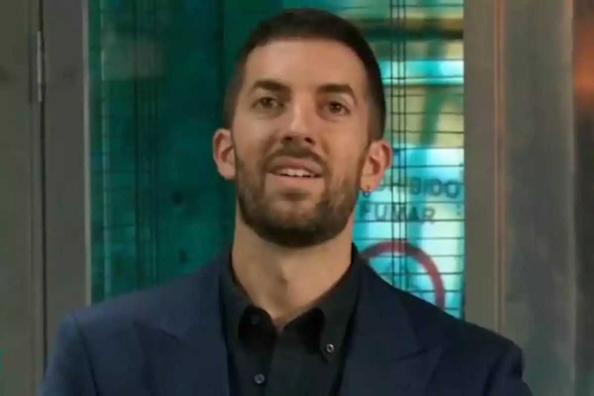 David Broncano de barba curta i cabell fosc porta una americana blau marí i una camisa negra davant d’un fons amb tons verds i blaus a La Revuelta.