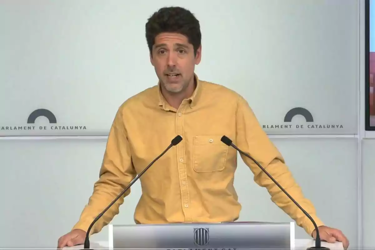 Un home amb camisa groga parla en un faristol amb el logotip del Parlament de Catalunya.