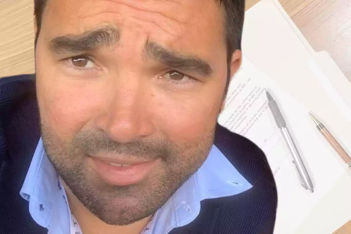 A man with a beard and expressive eyebrows looks at the camera, with documents and pens on a wooden table in the background.