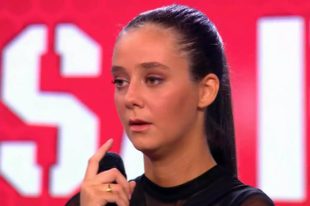 Victoria Federica con cabello oscuro recogido sostiene un micrófono frente a un fondo rojo en El Desafío.