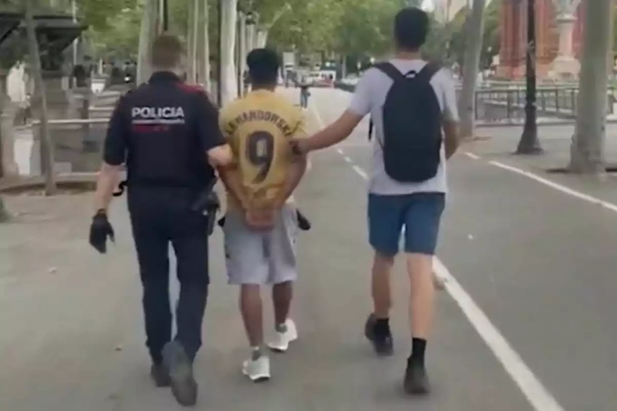 Un policia i una altra persona escorten un home amb una samarreta de futbol mentre caminen per un carrer arbrat.