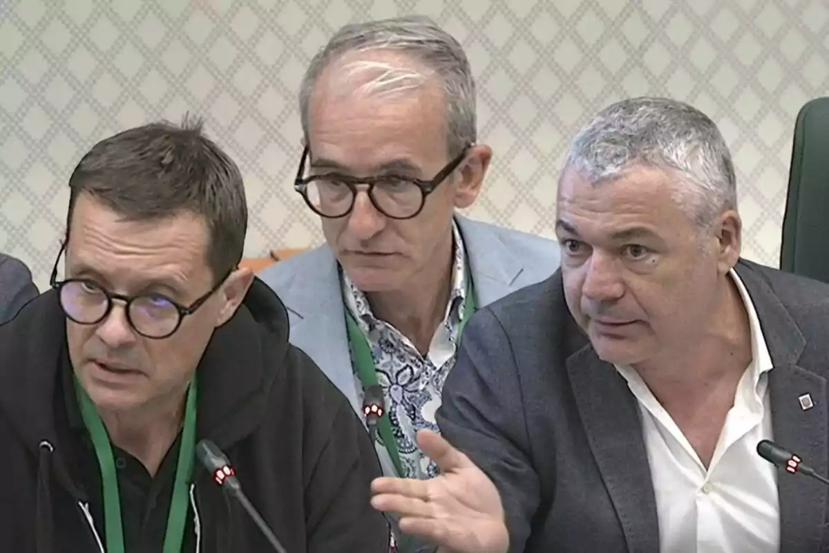 Tres homes asseguts en una taula de reunió amb micròfons i credencials verdes, conversant de manera seriosa.